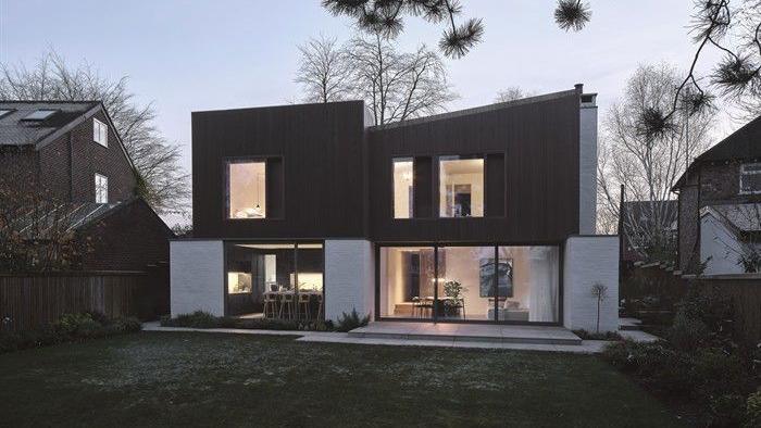 A modern house that looks like two square building pushed together. The top half of the exterior is dark brown, the bottom half is made up of large windows. The building on the left has a flat roof while the building on the right has a slight diagonal slant.