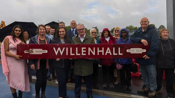 A large group of people holding a long red wooden sign. it says WILLENHALL in capital letters, with a British railways logo on either side. 