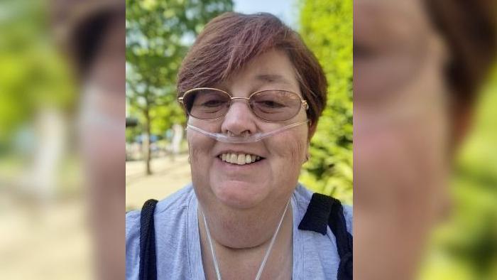 Sarah with a breathing tube in her nose smiling at the camera. She has short dark hair and is wearing a grey top. She is outside on a sunny day.