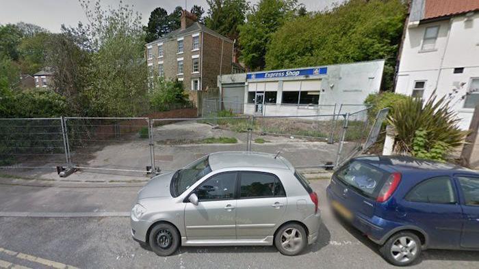Two cars parked by a fenced-off area of concrete. A building is behind the former forecourt with "Express Shop" written on it.