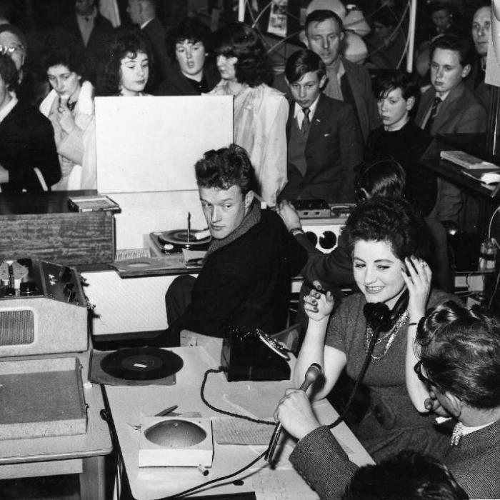 Black and white photo of the radio station with a man holding a microphone, a woman holding a phone receiver and other people in the background.