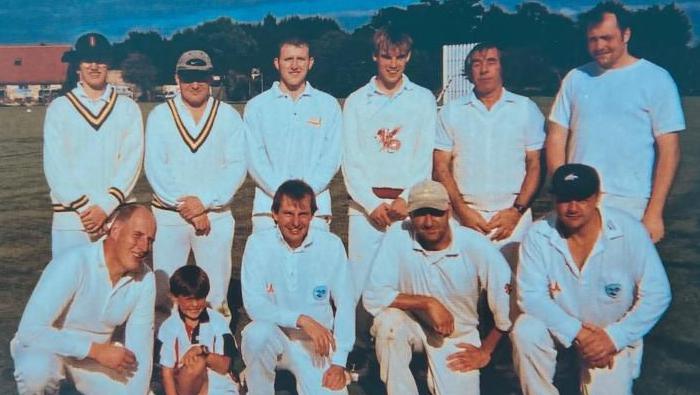 Ten men wearing cricket whites, and a small boy, in a team line-up on a cricket pitch. Six men are standing in the back row. Four men, and a boy, are crouching kneeling in the front row. Four of the men are wearing cricket caps.