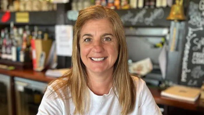 Sandrina Teece, a woman with long brown/ blonde hair, is pictured smiling. She is wearing a white T-shirt. There is a bar in the background.