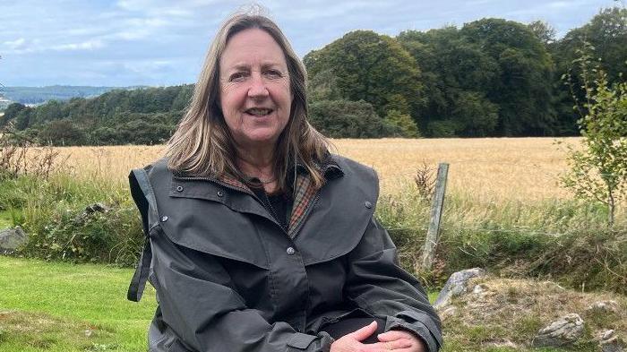 A woman sits in a garden bordered by a field of golden crops. She has shoulder-length blonde/brown hair. She is smiling a the camera and wearing a long green wax jacket. 