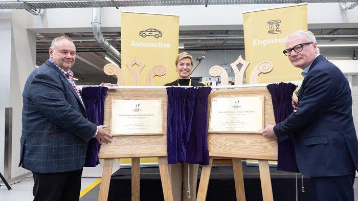 Two men are standing next to wooden boards with commemorative plaques on them, and purple curtains. A woman stands between them, smiling. There are yellow banners in the background