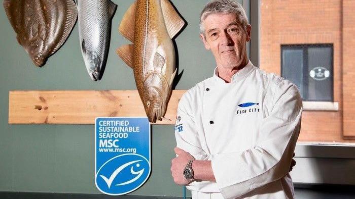 John Lavery stands in a white uniform with a blue logo that says fish city. He is smiling. Behind him there are three large fish on the wall and a blue certified sustainable seafood sign.