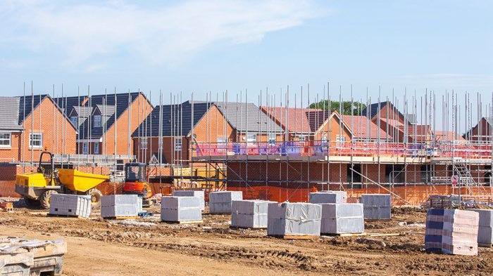 A newbuild estate with many red-brick houses, covered in scaffolding. An area of sand with a digger on it is in front of the houses.
