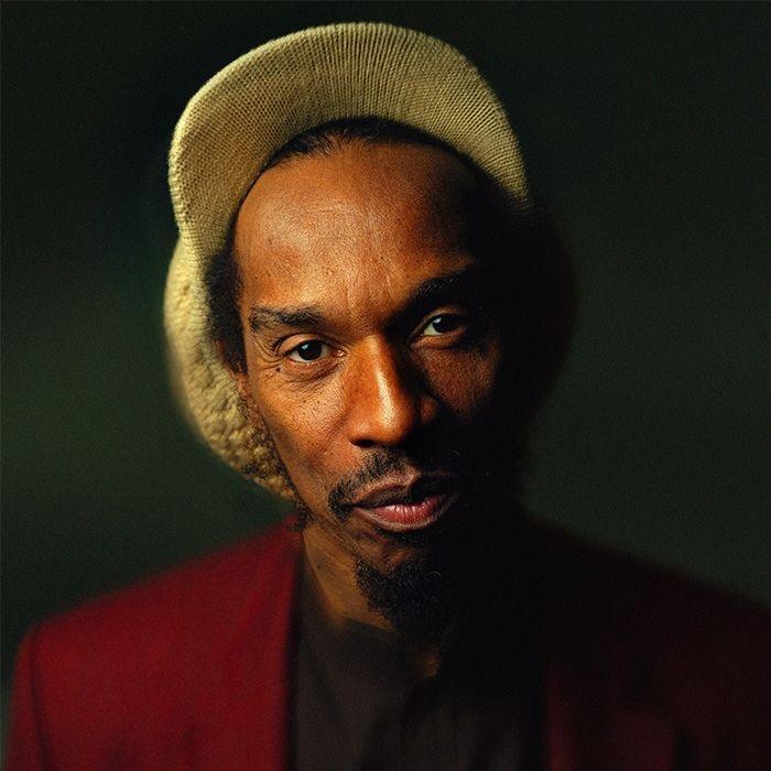 A photo portrait of Benjamin Zephaniah, looking straight at the camera and smiling slightly. He is wearing a large, cream woollen cap with a peak, and a red jacket.