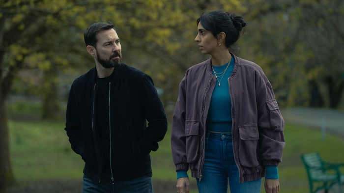 An image from TV show Fear with Martin Compston and Anjli Mohindra's characters in conversation while walking through a park - he is in a blue hoodie/jacket and she has a purple jacket, aqua top and blue jeans on