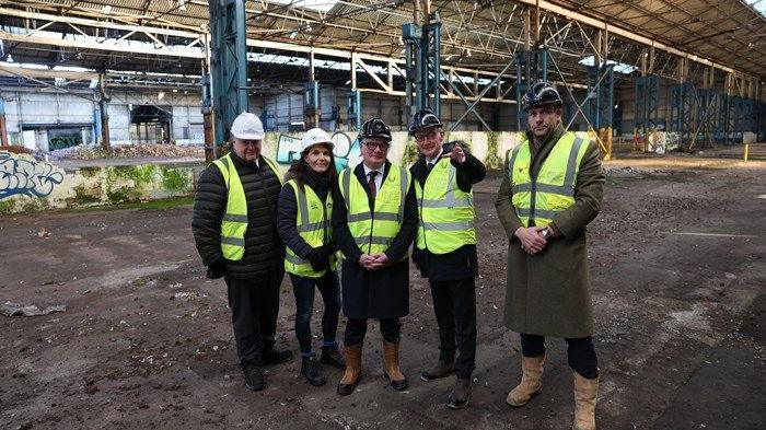 Four men and one woman wearing hi-viz jackets, boots and hard hats stand on a building site and smile at the camera