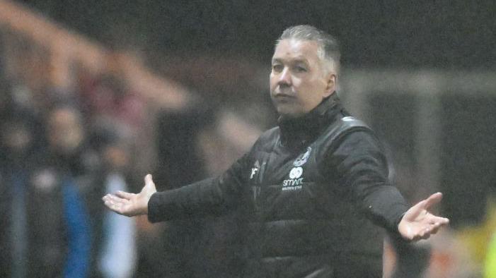 Darren Ferguson stood on the pitch during a match with his hands held out and a neutral expression on his face.