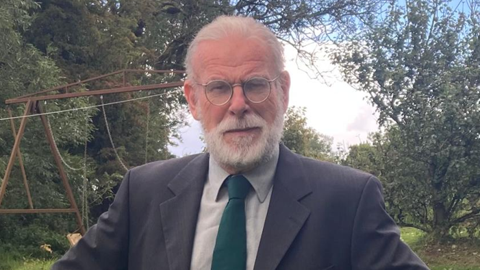 Shows a man with silver hair, silver beard and found glasses, with a grey jacket, light grey shirt and green tie, standing in front of trees