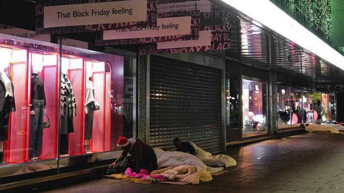 A high street lit up with shop windows. One window has four mannequins in pink booths and two people in sleeping bags can be seen outside. One is wearing a Santa hat. "That Black Friday feeling" is written on a shop sign above them.