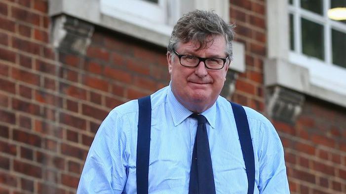 Crispin Odey wearing light blue shirt and dark blue tie and braces, standing outside a brown brick building