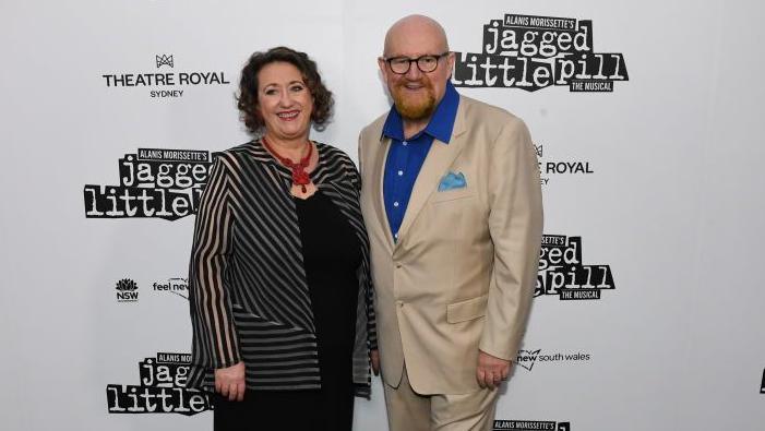 Dame Rosemary Squire is wearing a black dress and a black striped jacket and red necklace. Sir Howard Panter is wearing a cream suit and blue shirt. They are standing in front of a white wall with "Theatre Royal Sydney" and other signs in black lettering.