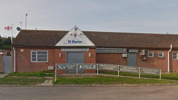 The clubhouse at FC Clacton, which is a one-storey building made of orange brick. It has a sign above a doorway that reads "welcome to FC Clacton". There are signs advertising the fact it sells, food, drinks and shows live sports on TV.