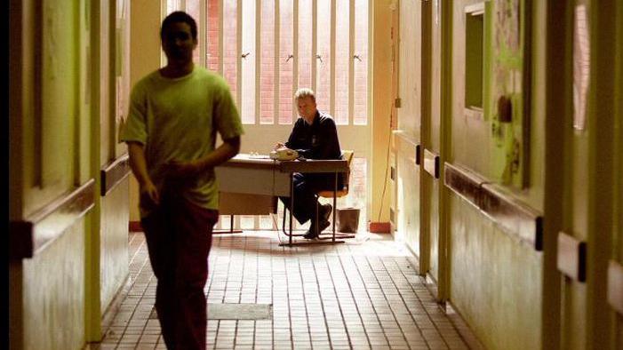 A young inmate walking along a prison corridor with a prison guard in the background