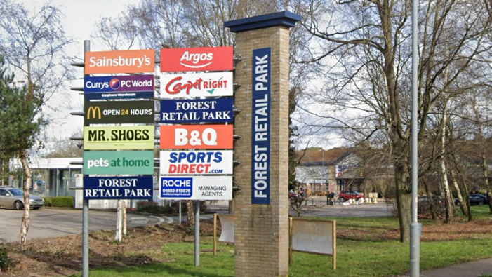 A sign is shown just off centre to the left, with the shops which are at Forest Retail Park in Thetford. In the background you can see parts of the retail complex. 