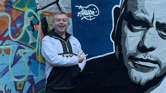 Mark Dallas next to a mural painted of himself dressed in a white The North Face quarter zip.