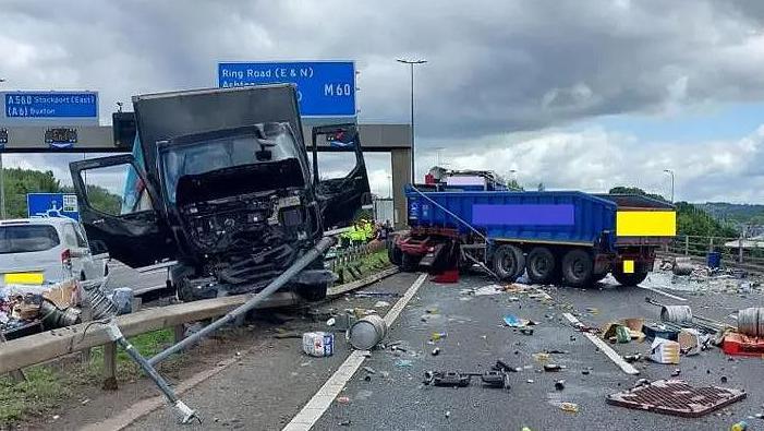 A lorry has crashed partially through a motorway central reservation barrier. There are metal barrels and debris all over the opposite carriageway. Another lorry has also crashed into the barrier on that side of the motorway.