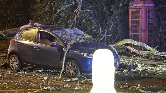 Debris from trees being blown over during Storm Darragh are spread across a road and a blue Ford car, which has had its windscreen smashed by some of the fallen branches. A red phone box is in the background.