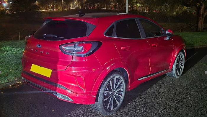 A red hatchback car is parked by the side of a road. The rear number plate has been blanked out.