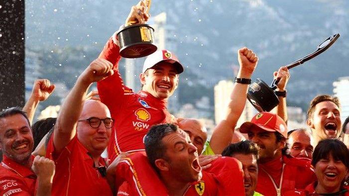 Charles Leclerc sits on the shoulders of a team member, holding a trophy aloft and grinning broadly. He is wearing a red Ferrari race suit and cap. He is surrounded by other jubilant colleagues, who are all wearing the red uniform of Ferrari and cheering.