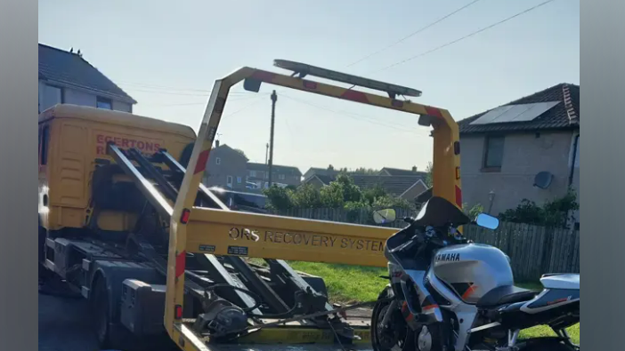 A Yamaha silver-and-black motorbike being loaded onto a pick-up truck on a housing estate on a sunny day.