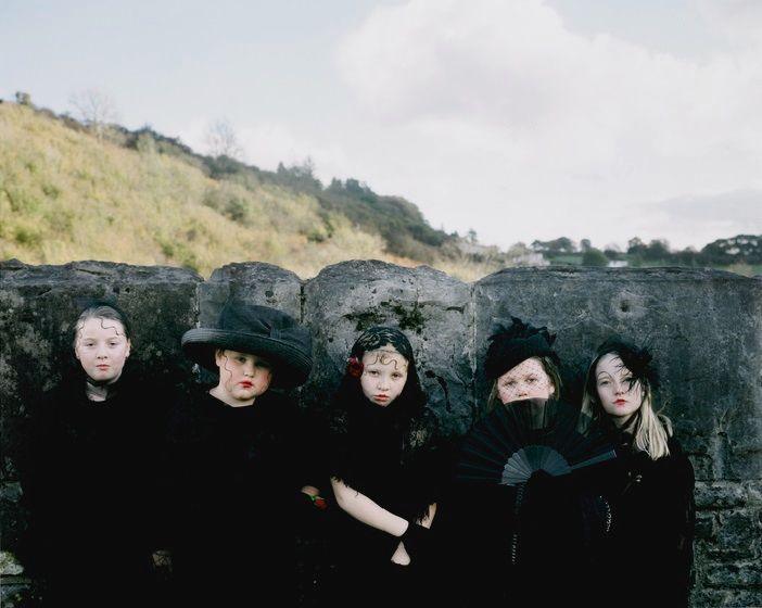 A photograph, It's Called Ffasiwn (Look It Up), taken in Merthyr 2016. Four young girls dresses in dramatic black dresses and hats or fascinators, standing in front of a stone wall with green hillside behind them.	