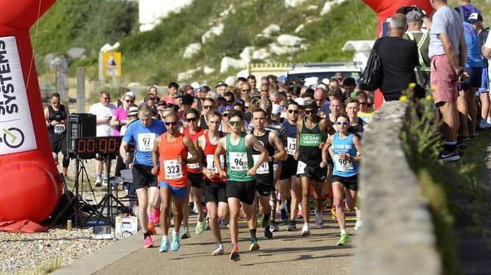 A large group of runners in vests and sunglasses run through an inflatable arch on a seafront path.