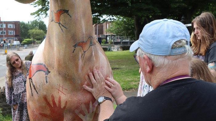 A man in a blue hat and a woman in a floral dress are placing their hands on a painted horse sculpture that they helped to design