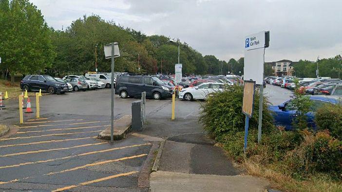 A car park with four long rows of parked cars. There are tall bushy trees in the background and to the left. There is a white parking sign to the right-hand side of the car park entrance. Also at the entrance, diagonal yellow line road markings lead to the parking area.