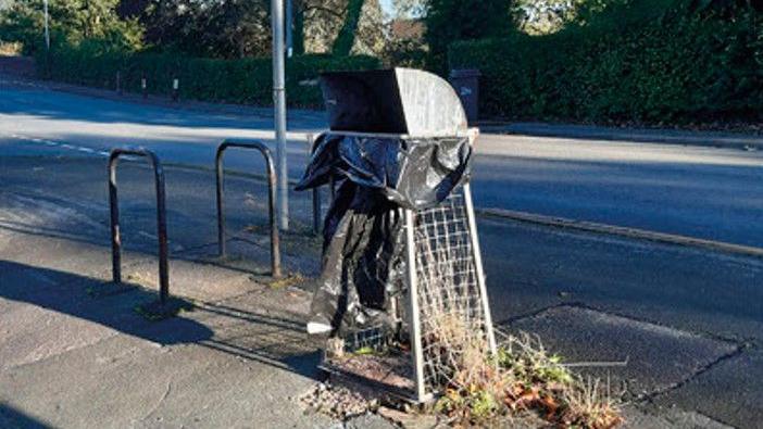 A mesh bin, with loose bin bag attached, at the side of a road, next to a pair of cycle racks. There is a pavement and a hedge in the background.