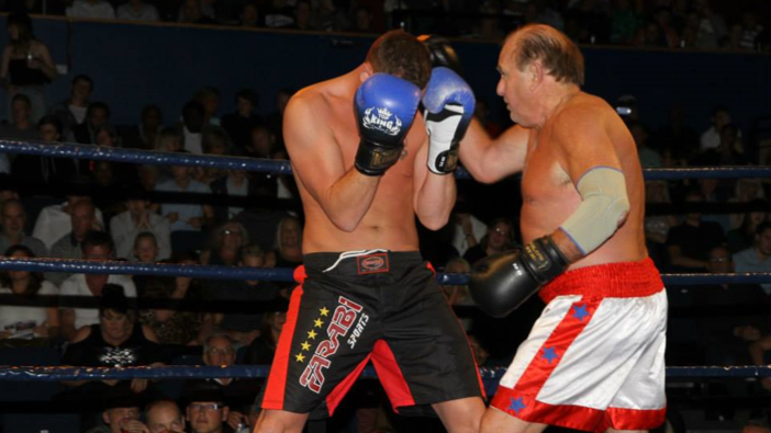 Two men boxing. On the left one with blue boxing gloves holding both of his hands in front of his head. On the right, an older man with dark hair. He is wearing white shorts with red stripes and blue stars. 