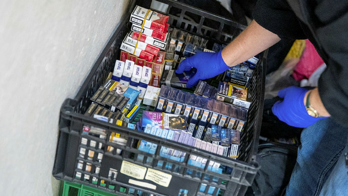 Crates of packets of cigarettes and tobacco. A person wearing blue gloves is sifting through the packets which are being held in a black crate which is stacked on top of a green crate. 