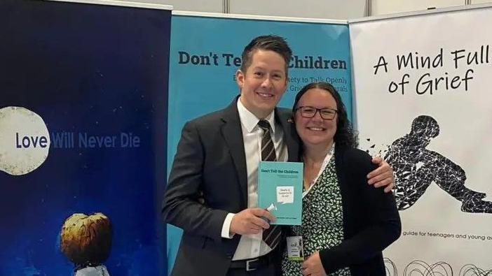 John Adams is in a black suit with a white shirt, and has his arm around Clare Shaw. She has brown hair and glasses and is wearing a floral dress. He is holding their book which is light blue