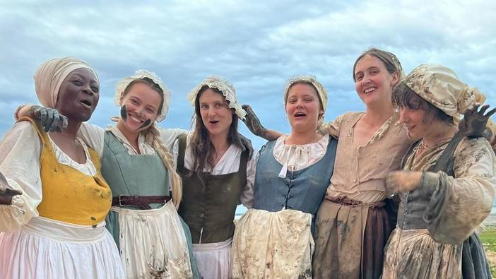 The Gossip actresses stand in a line, arm in arm, smiling at the camera. They wear dresses, aprons and hats in a 16th Century style. They are also covered in mud and dirt.