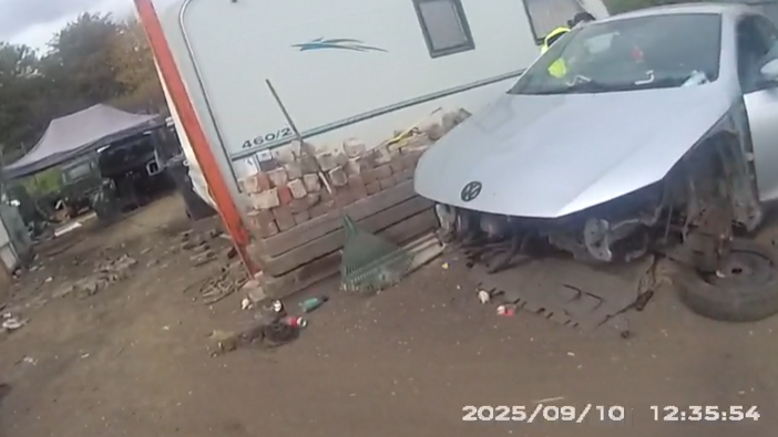 The image depicts an outdoor scene that appears to be part of a scrapyard or a vehicle dismantling area. On the right side, there is a partially dismantled silver Volkswagen car. The car is missing its front bumper and wheels, revealing its internal components. Behind the car, a white trailer is visible with the number "460/2" written on it.