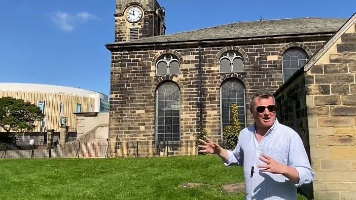 Luan Hanratty. He is wearing a blue shirt and sunglasses and is standing outside an old building, with a clock tower.