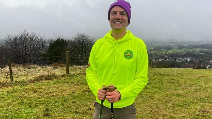 A man wearing a neon green hoodie and purple beanie hat, and brown trousers, smiles at the camera as he holds two black walking sticks, and stands in a green field, with rolling fields as a view behind him. It is a cloudy, foggy day