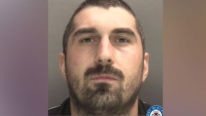 A police picture of a man looking at the camera. He has brown cropped hair, a moustache and beard.
