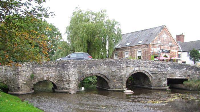 A stone bridge with a car driving over it. Underneath the bridge is a river. There are green trees behind the bridge.