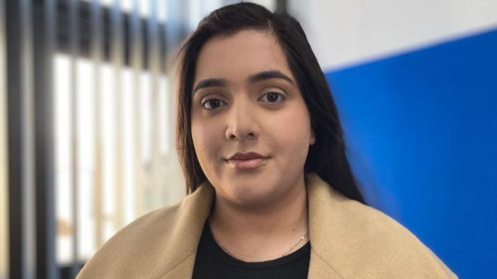 Marium Zameer, wearing a beige coat and black top, standing in front of a blue wall and, to her left, a window with blinds open.