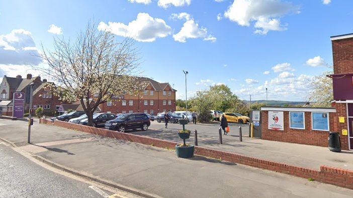 The image shows a car park next sandwiched between buildings. Several trees are growing in the car park, beginning to show leaves. A purple sign with a silver moon and the words "Premier Inn" can be seen in the left corner.