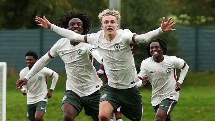 Jesse Derry, wearing Chelsea's light-coloured away kit, has his arms outstretched as he celebrates a goal in an academy game, as his team-mates run to keep up with him