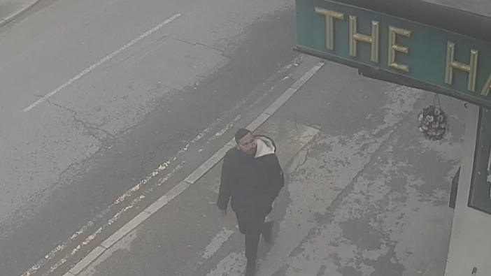 A CCTV image in black and white showing a man with a close-cropped beard and short hair walking along the pavement beside a main road.