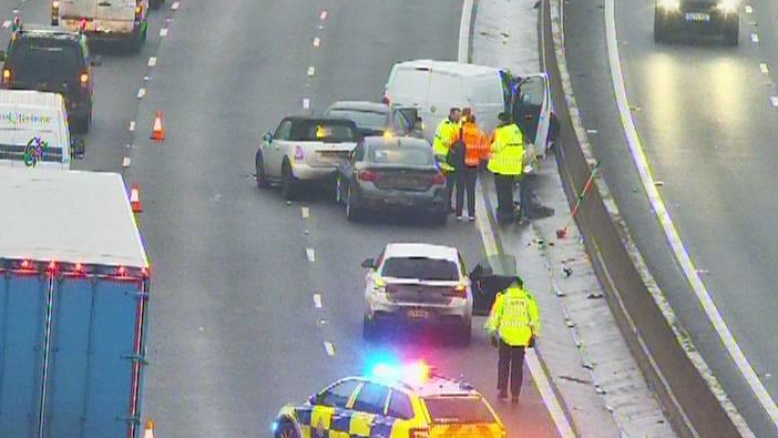 Traffic passes by a a multi-vehicle crash. People in hi-visibility jackets stand near the crash site