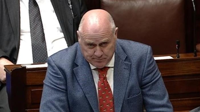 Noel Graelish speaking in the Dáil. He is bald and wearing a blue blazer, white shirt and red tie. The brown leather Dáil seats can be seen in the background with two other TDs sitting in them. 