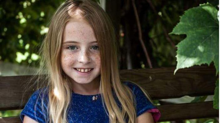 Cimarron Thomas  is staring into the camera. She has dark blonde hair and blue eyes. She is wearing a blue dress with short sleeves. She is sitting on a bench with trees in the background