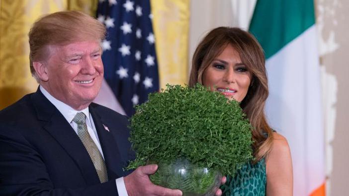 Donald Trump and his wife Melania Trump posing with a bowl of shamrock at the White House in 2018.  Trump is wearing a navy suit, white shirt and green tie and Mrs Trump is wearing a green animal print dress.  The Irish and US flags are behind the couple.